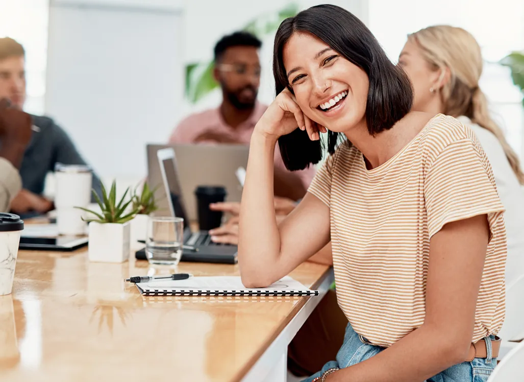 Colaboradora sorrindo na mesa de trabalho, com a equipe ao fundo